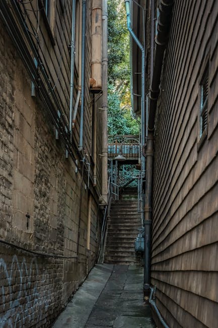 A narrow alleyway between two buildings, with the left side constructed from aged brick featuring some graffiti and multiple black and grey metal pipes running vertically and horizontally along the wa
