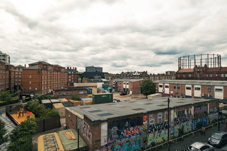 A young man with a serious expression, wearing a black cap and a white polo shirt, stands inside an open van that is filled with neatly stacked cardboard boxes of various sizes, some secured with plas